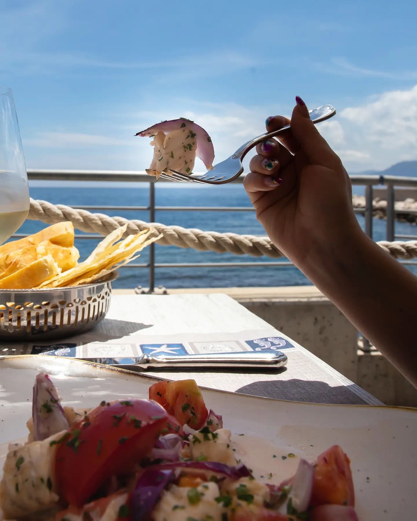 Forchetta con pesce crudo e cipolla rossa sopra un piatto di pomodori, terrazza con corda e vista sul mare azzurro