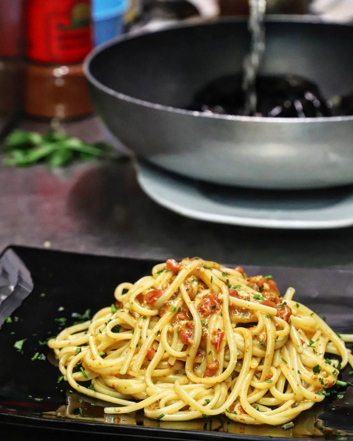 Spaghetti con sugo di mare, pomodorini e prezzemolo su piatto nero, con padella antiaderente sullo sfondo, Il Pescatore